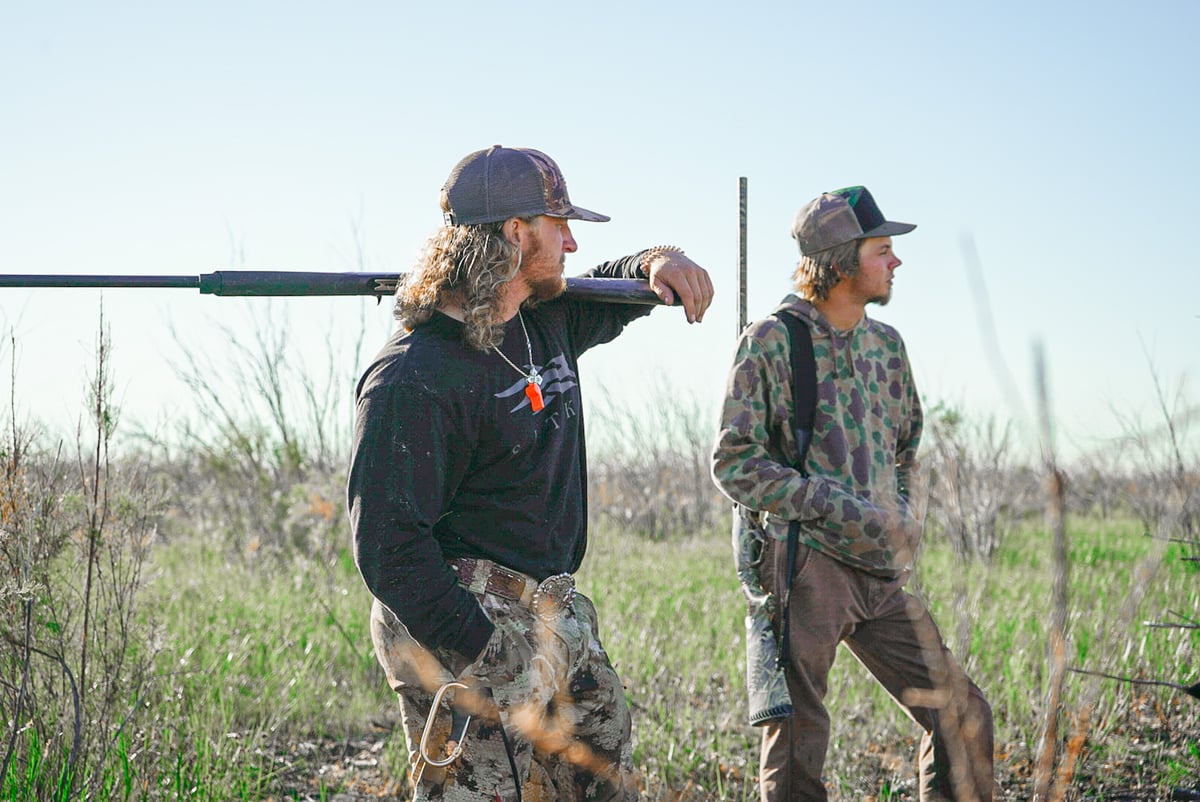 Two male hunters looking off into the distance, one holding a rifle.